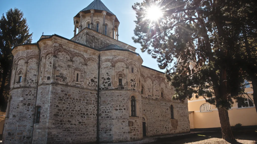 Kamena crkva manastira Novo Hopovo sa visokim kubetom okruženim stubićima, pored nje četinar i žuti konak