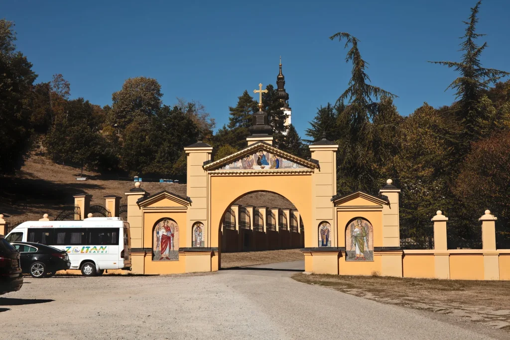 Žuta ulazna kapija manastira Grgeteg, pored nje parkiran turistički minibus i automobili