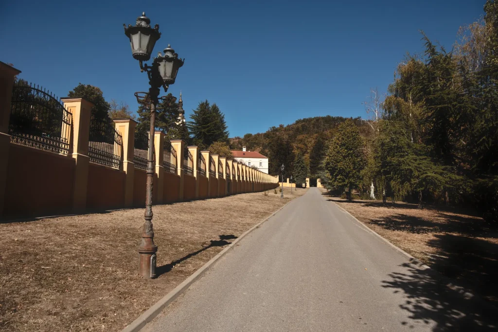 Asfaltni put uz dugi žuti ogradni zid sa kovanom ogradom i starinskom lampom, od kapije ka manastiru Grgeteg