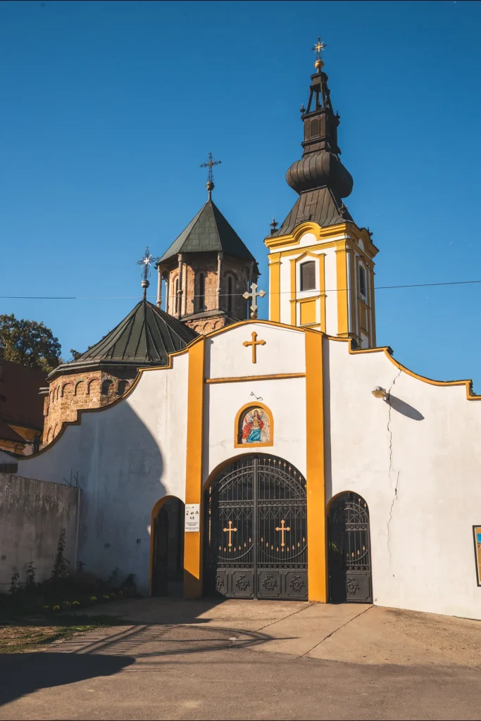 Bela ulazna kapija manastira Privina Glava sa žutim pilastrima, ikonom i kovanim vratima, iza nje kube crkve i zvonik